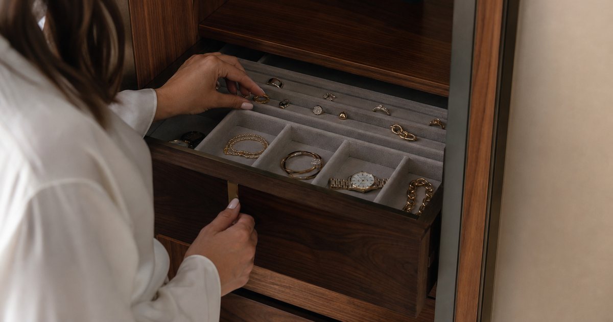 Open velvet-lined jewelry drawer in a walnut dressing room cabinet showing compartments with rings, bracelets, and a watch, a woman's hand reaching in, soft directional lighting from above