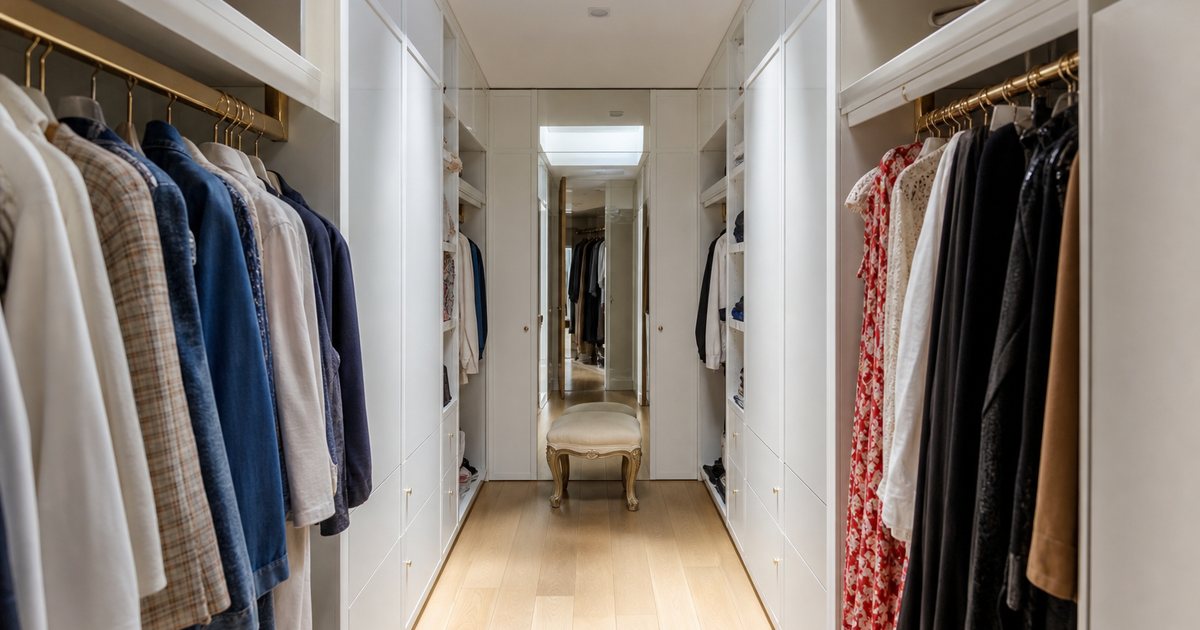 A compact but elegant walk-in closet with floor-to-ceiling white lacquered wardrobes on both sides of a narrow aisle, a full-height mirror at the far end, brass rods, and a small upholstered stool, lit by a skylight